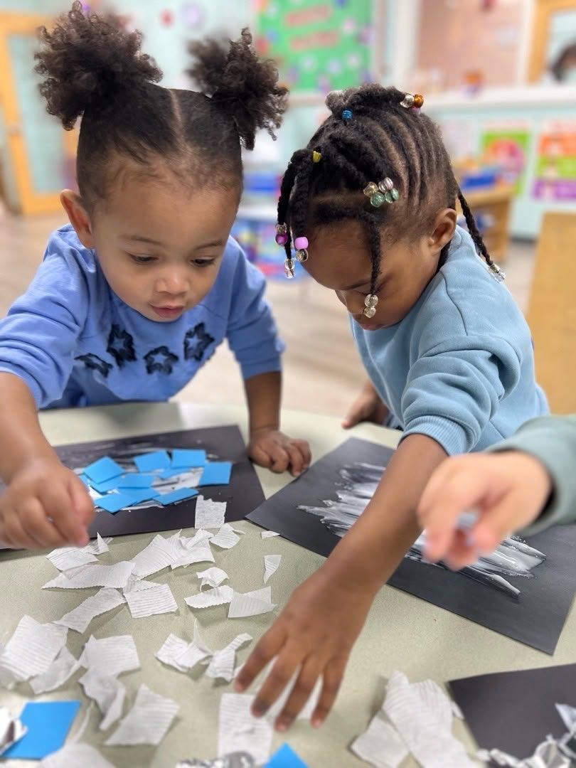 Two children cutting and arranging paper shapes at a classroom table.