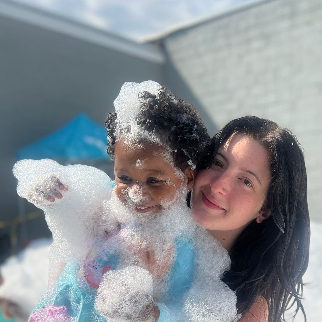 A teacher holding a laughing child, both covered in soap foam during outdoor play at ESPIN.