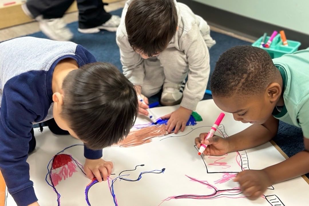 Four children drawing side by side on one long roll of paper.
