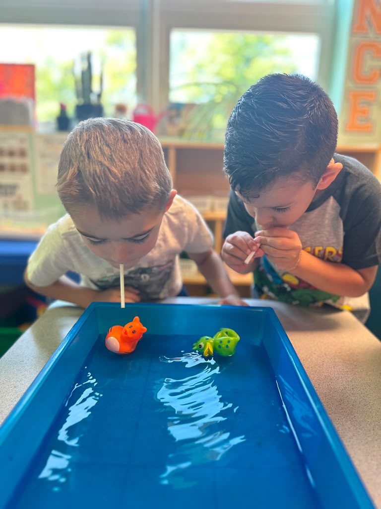 Two children blowing toy boats across a classroom water table.