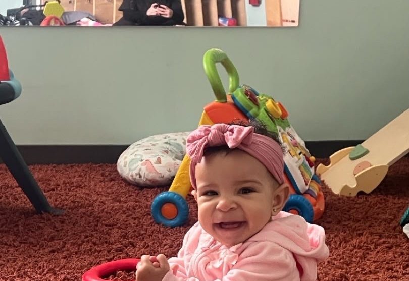 An ESPIN infant sitting on the classroom rug, smiling among soft toys.