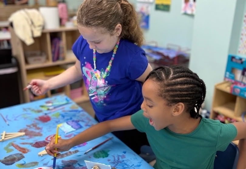 Two preschool children painting side by side at a classroom art table.